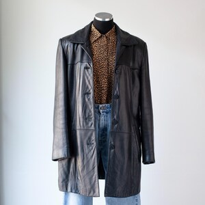 May include: A black leather coat with a button-up front, paired with a leopard print button-down shirt and blue jeans. The coat is knee-length and has a classic, tailored design. The outfit is displayed on a mannequin.