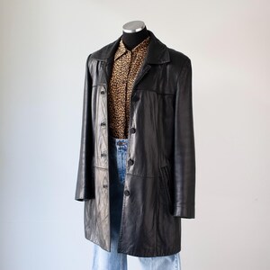 May include: A black leather coat with a button-up front, worn over a leopard print button-down shirt and blue jeans. The coat has a classic, tailored design and falls to mid-thigh length. The outfit is displayed on a mannequin.