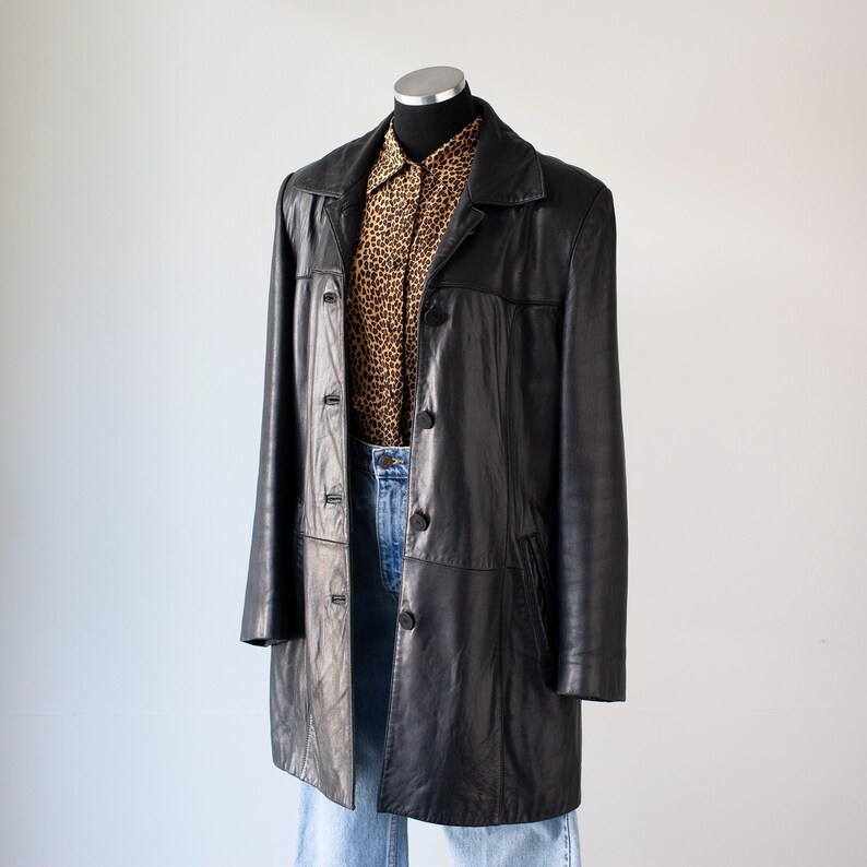 May include: A black leather coat with a button-up front, worn over a leopard print button-down shirt and blue jeans. The coat has a classic, tailored design and falls to mid-thigh length. The outfit is displayed on a mannequin.
