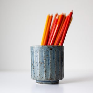 May include: A blue ceramic pencil holder with vertical grooves, filled with red and yellow pencils. The pencils are arranged upright in the holder, creating a colorful display. The holder has a textured surface and a small base.