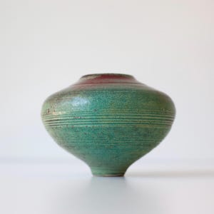 May include: A handcrafted ceramic vase with a rounded body and a narrow base. The vase features horizontal textured lines and a gradient of colors, transitioning from a deep red at the top to a vibrant green in the middle, and a lighter green at the bottom.