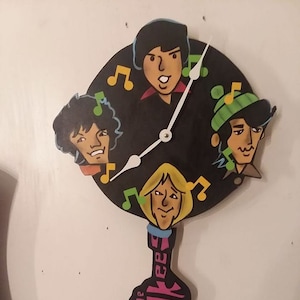 May include: A black and white clock with a pink guitar-shaped base. The clock face features four cartoon faces of the band The Monkees, with musical notes around the edge. The clock hands are white.