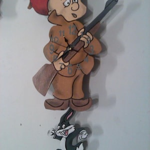 May include: A wooden wall clock featuring a cartoon character dressed as a hunter holding a rifle. The clock face has Roman numerals and the character is standing on top of a cartoon rabbit running away.