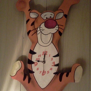 May include: Orange and black cartoon tiger clock with a white face and black hands. The clock is hanging on a wood panel wall.