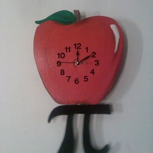 May include: A red apple-shaped clock with a black face and black hands. The clock is hanging from a black pi symbol.