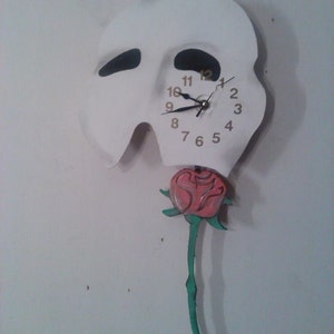 以下が含まれることがあります： 黒い時計の針と金の数字が付いた、オペラ座の怪人のマスクの形をした白い時計。赤いバラが緑の茎で時計の下からぶら下がっている。