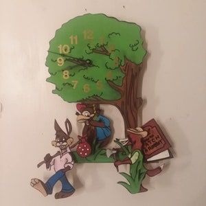 May include: A wooden wall clock shaped like a tree with a squirrel, rabbit, and fox. The clock face has gold numbers and hands. The squirrel is holding a bag with red polka dots. The fox is carrying a book that says "How to Catch a Rabbit".