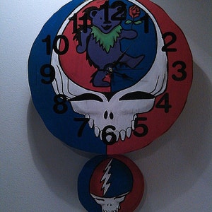 May include: A round clock with a red, white, and blue design. The clock face features a skull with teeth and a bear with a flower. The clock hands are black. The clock is hanging on a white wall. The clock has a smaller, round, red, white, and blue pendant with a skull and lightning bolt design.