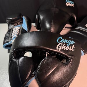 Könnte beinhalten: Schwarzes Box-Set mit Kopfschutz und Handschuhen. Der Kopfschutz und die Handschuhe haben den Text "Congo Ghost" in Hellblau gestickt. Die Handschuhe sind 10 oz und haben weiße Schnürsenkel. Die Innenseite ist hellblau.