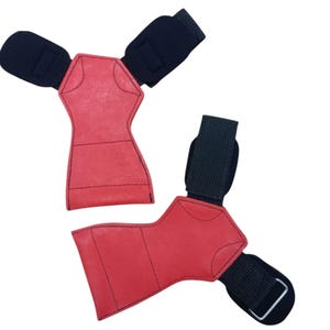 May include: Two red weightlifting grips with black wrist straps. The grips are made of a textured material and have a unique shape. The wrist straps have a hook and loop closure.