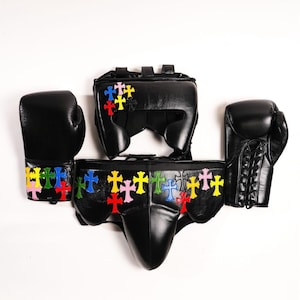 May include: Black boxing gear set including gloves, headgear, and a protective cup. Each piece is adorned with colorful cross designs. The gloves have lace-up closures. The set is displayed against a white background.