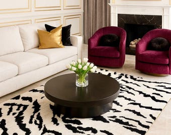 May include: A living room with a white sofa, gold and black throw pillows, and two burgundy armchairs. A black and white patterned rug sits beneath a dark brown coffee table with a vase of white flowers. The walls are white with gold trim.