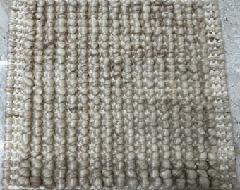 Puede incluir: Muestra de alfombra cuadrada y texturizada con una paleta de colores neutros. La alfombra presenta un diseño de pelo en bucle en tonos beige y marrón, con un borde blanco. La vista de cerca resalta la construcción tejida y las variaciones del hilo.