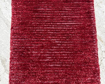 Puede incluir: Una alfombra rectangular y afelpada de color burdeos intenso. La alfombra tiene una superficie texturizada y peluda con líneas horizontales, creando una apariencia suave y acogedora. La alfombra está diseñada para uso en interiores.