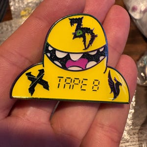 May include: A yellow enamel pin featuring a stylised character with a wide, toothy grin, a purple mouth, and a green eye. The design includes a black symbol on the left, the text TAPE 8 in the centre, and a black bird-like silhouette on the right.