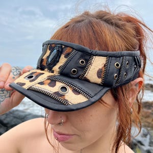 Könnte beinhalten: Schwarzes Leder-Visier mit einem Leopardenmuster-Panel und silbernen Ösen. Das Visier hat einen schwarzen Stein und Reißverschlussdetails. Ein einzigartiges Accessoire für den Sonnenschutz.