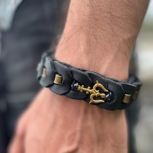 Puede incluir: Pulsera de cuero trenzado negro con detalles de latón y un dije de tridente dorado. La pulsera mide aproximadamente 2 cm de ancho y tiene un cierre seguro. Un accesorio elegante para el uso diario.