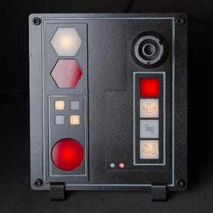 May include: A black control panel with a variety of buttons and lights. The panel has a red light, a white light, a red button, a white button, and a black button. The panel also has a small black speaker.