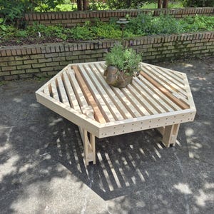 Könnte beinhalten: Ein helles, sechseckiges Couchtisch aus Holz mit einer Lamellenplatte. Der Tisch hat vier Beine und trägt einen kleinen dekorativen Topf mit Grünzeug. Der Tisch ist etwa 91 cm breit.