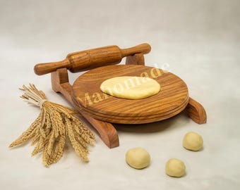 Puede incluir: Una tabla circular de madera marrón con un rodillo a juego sobre un soporte. Un trozo de masa de color amarillo pálido descansa sobre la tabla, junto a tres pequeñas bolas de masa. Un manojo de tallos de trigo seco se sitúa a la izquierda.