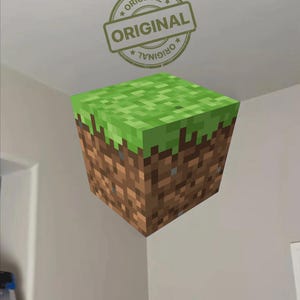 Puede incluir: Un cubo pixelado, que recuerda a un bloque de videojuego, con una parte superior verde y lados de tierra marrón. La palabra "ORIGINAL" está estampada encima del cubo en un diseño circular.