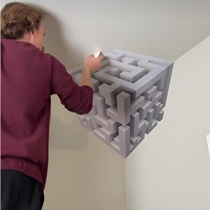 May include: A wall decal featuring a three-dimensional cube design with a complex maze pattern in shades of gray and white. The graphic creates an optical illusion of depth, appearing as a geometric structure protruding from the corner of a wall.