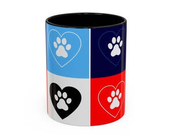 Könnte beinhalten: Eine Keramik-Kaffeetasse mit schwarzem Innenraum und farbenfrohem Außendesign. Die Außenseite ist in vier Abschnitte unterteilt: Hellblau, Marine, Weiß und Rot. Jeder Abschnitt zeigt eine Herzform mit einem Pfotenabdruck.