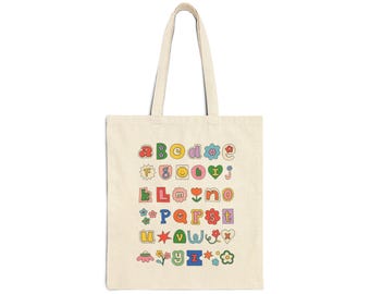 May include: A cream-colored canvas tote bag featuring a colorful alphabet print. Each letter is displayed in a unique, playful font style, interspersed with small graphic icons including flowers, stars, hearts, and a smiley face in various bright colors.