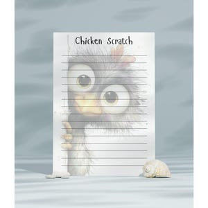 May include: A notepad with the text "Chicken Scratch" features a cartoon chicken peeking from behind the paper. The chicken has large eyes and a fluffy appearance. A seashell and a small stone are in the foreground.