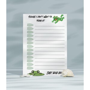 May include: A white notepad with horizontal lines and a green frog illustration. The notepad has the text "THINGS I DON'T WANT TO FROG-ET" and "JUST RIB-BIT". A small white seashell and a white stone are next to the notepad.