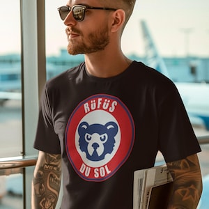 May include: A black short-sleeved t-shirt featuring a circular graphic on the chest. The graphic contains a stylized blue bear head centered within a red ring, surrounded by the white text RÜFÜS DU SOL. The shirt has a crew neckline.