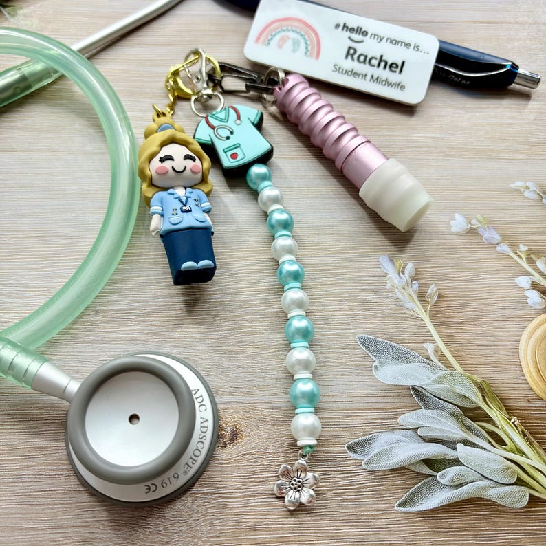 May include: A collection of medical-themed accessories, including a stethoscope, a keychain with a nurse figurine and scrubs charm, a pen, and a name tag that reads "Rachel Student Midwife". The keychain features a beaded strand with pearl and turquoise beads.