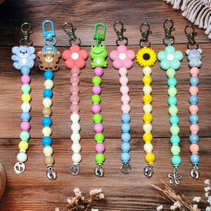 May include: A collection of colorful beaded pacifier clips with various charms, including a teddy bear, monkey, flower, and frog. Each clip features a metal clasp and a charm at the bottom. The beads are in a rainbow of colors.