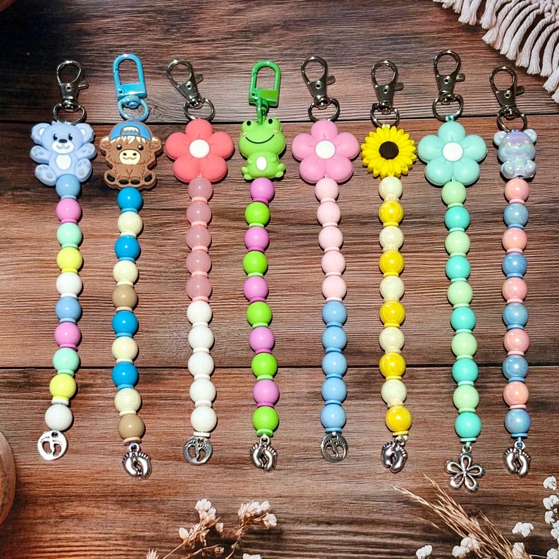 May include: A collection of colorful beaded pacifier clips with various charms, including a teddy bear, monkey, flower, and frog. Each clip features a metal clasp and a charm at the bottom. The beads are in a rainbow of colors.