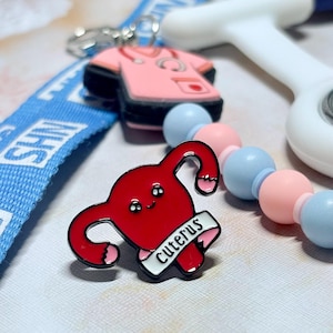 May include: A red enamel pin in the shape of a uterus with the word "Cuterus" on a banner. The pin has a black outline and pink accents. Other items in the image include a pink scrubs charm, a blue lanyard, and a beaded necklace.