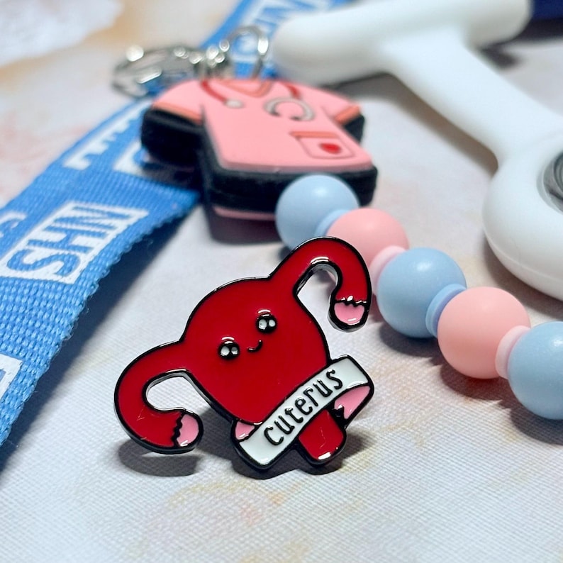May include: A red enamel pin in the shape of a uterus with the word "Cuterus" on a banner. The pin has a black outline and pink accents. Other items in the image include a pink scrubs charm, a blue lanyard, and a beaded necklace.