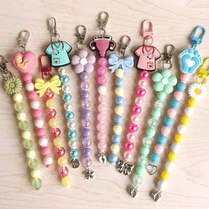 May include: A collection of beaded keychains in various pastel colors. Each keychain features a unique charm, including a flower, bow, scrubs, and a baby in a heart. The keychains are attached to silver-tone clasps.