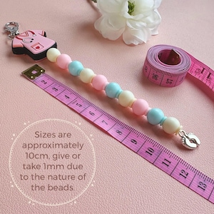 May include: A baby pacifier clip with pastel-colored beads, a medical scrubs charm, and a silver-colored foot charm. The clip is approximately 4 inches long, with the text "Sizes are approximately 10cm, give or take 1mm due to the nature of the beads."