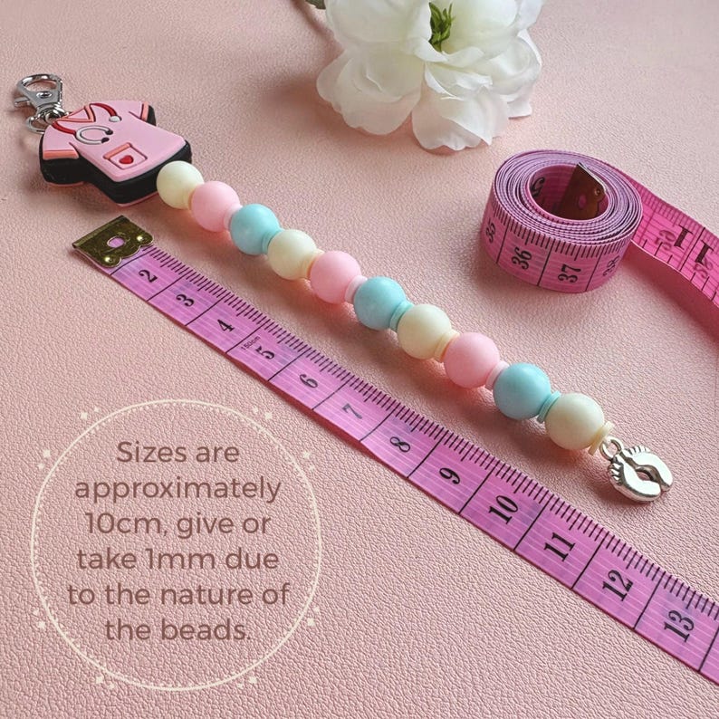 May include: A baby pacifier clip with pastel-colored beads, a medical scrubs charm, and a silver-colored foot charm. The clip is approximately 4 inches long, with the text "Sizes are approximately 10cm, give or take 1mm due to the nature of the beads."