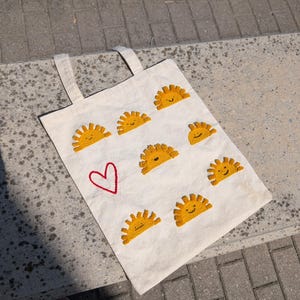 May include: Off-white canvas tote bag featuring embroidered yellow suns and a red heart. The bag has two handles and is designed for carrying items. The suns have smiling faces, adding a cheerful touch to the design.