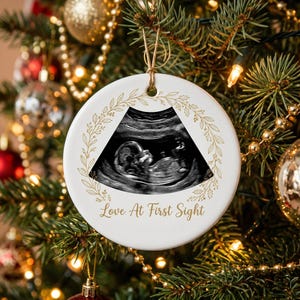 May include: A round white ceramic ornament featuring a black and white ultrasound image of a fetus. The design includes a gold-colored botanical wreath border and the text "Love At First Sight" printed in gold script below the ultrasound image.
