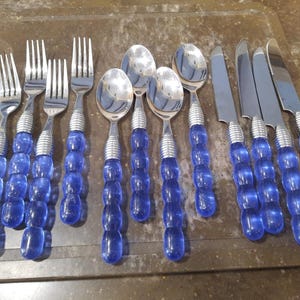 Puede incluir: Un juego de cubiertos de acero inoxidable con mangos de cuentas azules. El juego incluye tenedores, cucharas y cuchillos. Los mangos son de color azul vibrante y los cubiertos están colocados sobre una superficie marrón.