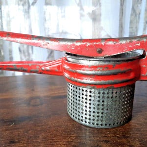 Puede incluir: Prensa puré de patatas vintage roja y plateada. La herramienta de cocina tiene una cesta de metal perforada y un mango rojo. La pintura está desgastada, lo que le da un aspecto rústico. Está diseñada para machacar patatas y otros alimentos blandos.