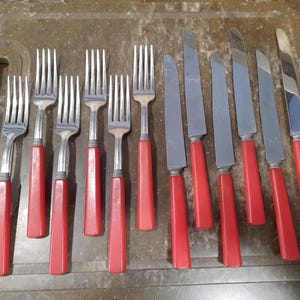 May include: A collection of silverware, including forks and knives, with red handles. The forks have four tines, and the knives have a smooth, silver blade. The silverware is arranged on a brown surface.