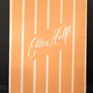 Puede incluir: Una caja rectangular con fondo melocotón y rayas blancas verticales. El texto en cursiva "Ellen Hall" está centrado en la caja. La caja tiene un diseño sencillo y limpio.