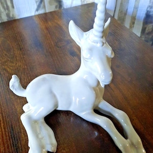 May include: A white ceramic unicorn figurine in a reclining pose. The mythical creature has a spiraled horn, pointed ears, and a small beard. The figurine is on a dark wooden surface.
