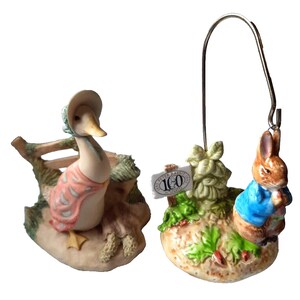 May include: Two ceramic figurines featuring characters from Beatrix Potter's stories. One depicts a duck in a bonnet and pink shawl, the other a rabbit in a blue jacket. A sign reads "Peter Rabbit 100 Limited".