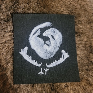 Op de afbeelding: Vierkante zwarte patch met een witte illustratie van twee verstrengelde dieren, een schedel- en tandontwerp eronder en een klein symbool. De achtergrond is een bruin, getextureerd oppervlak.