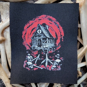 May include: Black square patch with a white and red illustration of a spooky house with a red sun-like background. The house is perched on a claw-like structure with skulls. The patch is surrounded by bones.
