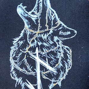 Op de afbeelding: Zwarte stof met een witte en gouden illustratie van een huilende wolfskop. De bek van de wolf is open, met tanden zichtbaar. Een kruis staat onder de kop van de wolf, en een gouden lijn omcirkelt zijn nek.
